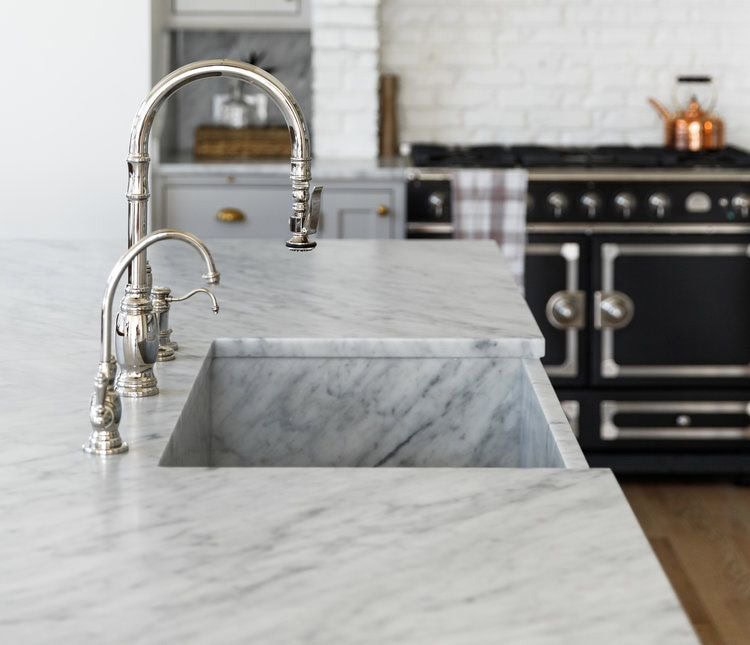 A modern kitchen with a marble countertop featuring two sleek, silver faucets. The backsplash is white brick, and in the background, there's a vintage-style black oven with copper pots on top.