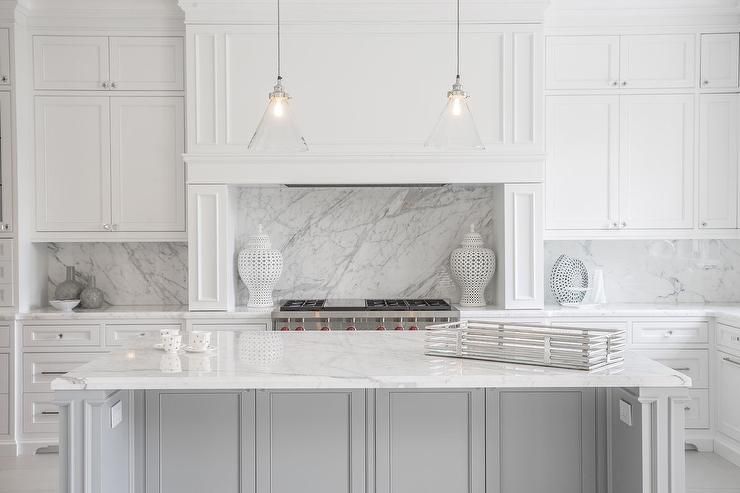 A modern kitchen with white cabinets and marble countertops. Two pendant lights hang above the island, which has decorative items. A stove is built into the countertop, and ornate vases add elegance to the space.