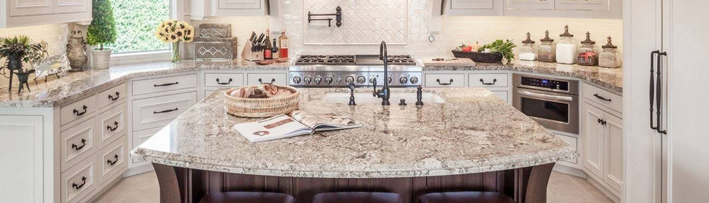 Modern kitchen with granite countertops, a large island, and stainless steel appliances. White cabinets line the walls, and a variety of decorative items, including a basket and cookbook, are on the island. The room is well-lit with natural light.