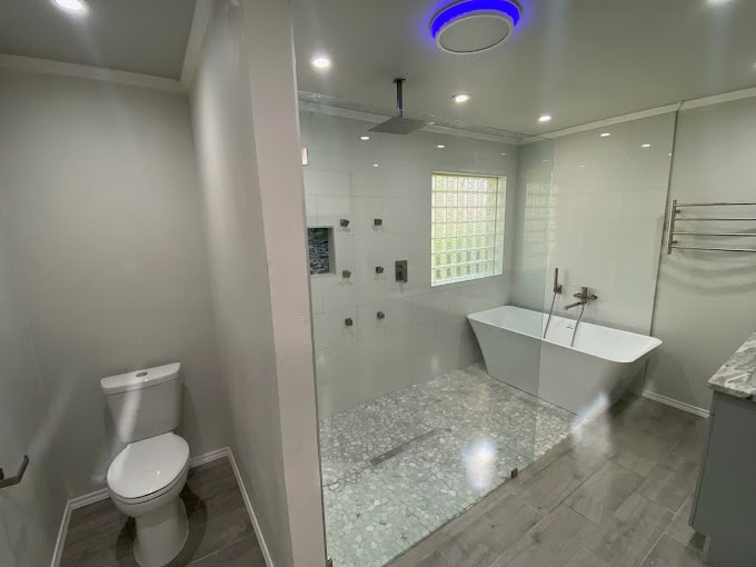 Modern bathroom with a glass-enclosed shower featuring a rainfall showerhead and pebble-style floor. A freestanding bathtub is positioned beside a window. The toilet is separated by a half wall. The space has sleek gray and white decor.