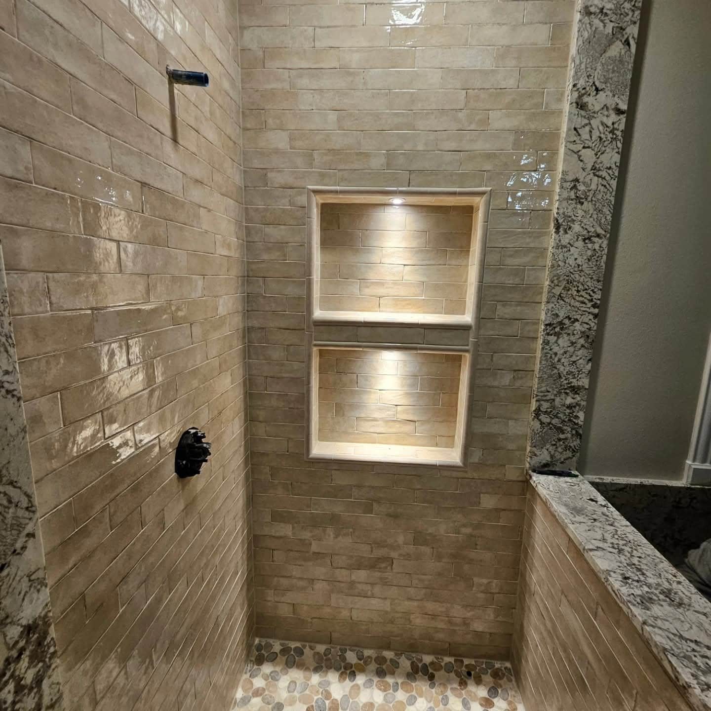 A modern shower with beige, rectangular tiles on the walls and pebble-style flooring. It features a black showerhead and a built-in niche with two shelves, each illuminated by a small light.