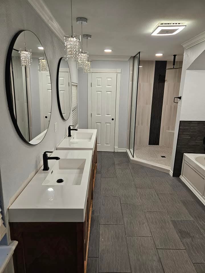 Modern bathroom featuring two sinks with black faucets, oval mirrors, and hanging crystal lights. A shower with glass doors is on the right, next to a bathtub. The floor is tiled in gray, and there is a white door in the background.