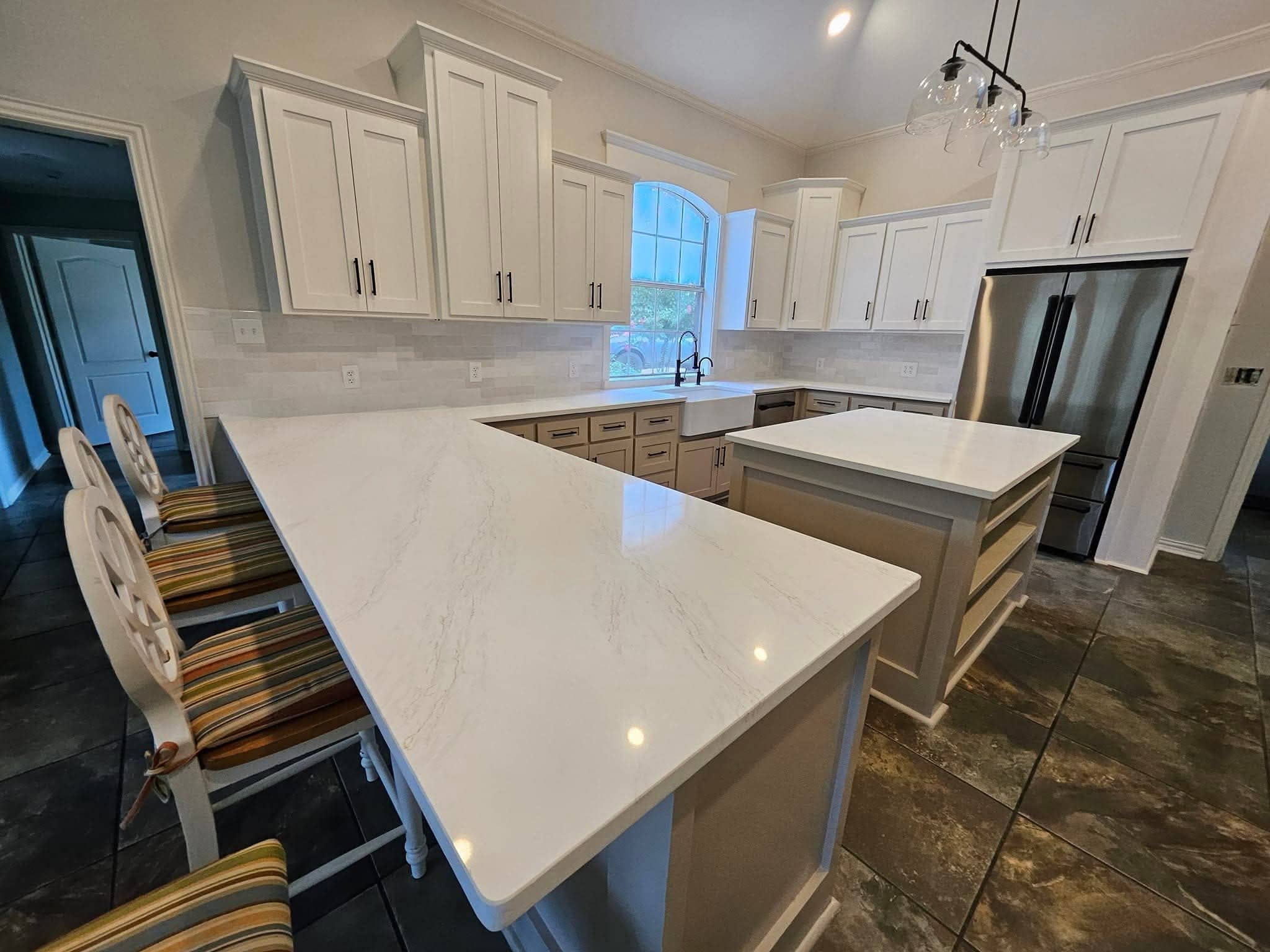 A spacious kitchen with white cabinets, marble countertops, and a large island. There are three striped barstools at the island. Stainless steel appliances are visible, and a large window provides natural light. The floor is tiled in a dark pattern.