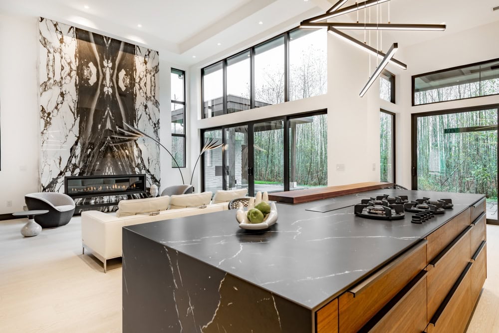 A modern living room with high ceilings and large windows, featuring a striking marble fireplace wall. The space includes a sleek black kitchen island with a gas cooktop, wooden cabinetry, and stylish white sofas facing the fireplace.