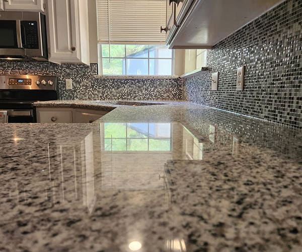 Close-up of a clean, polished granite kitchen countertop with a mosaic tile backsplash, white cabinets, and a window reflecting natural light onto the surface—perfectly complementing expert flooring installation throughout the space.
