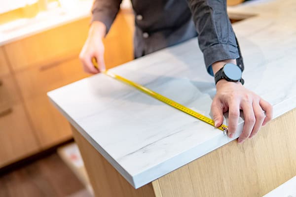 A person measures the width of a white marble kitchen countertop with a yellow tape measure, preparing for flooring installation. The surrounding cabinetry is wooden, and the person wears a dark long-sleeve shirt and a watch.