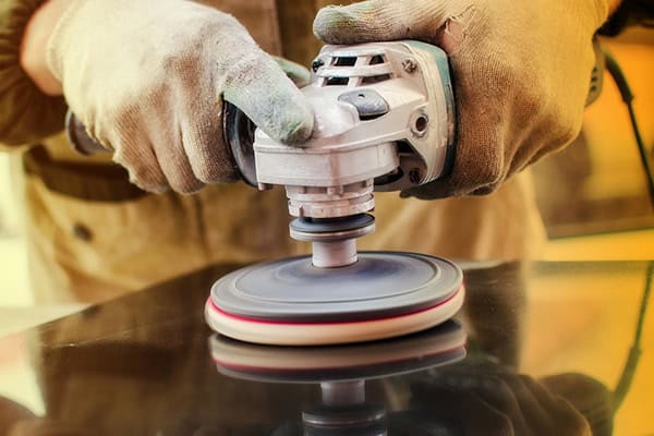 A person wearing protective gloves uses a power tool with a spinning disc to polish or buff a reflective black surface.